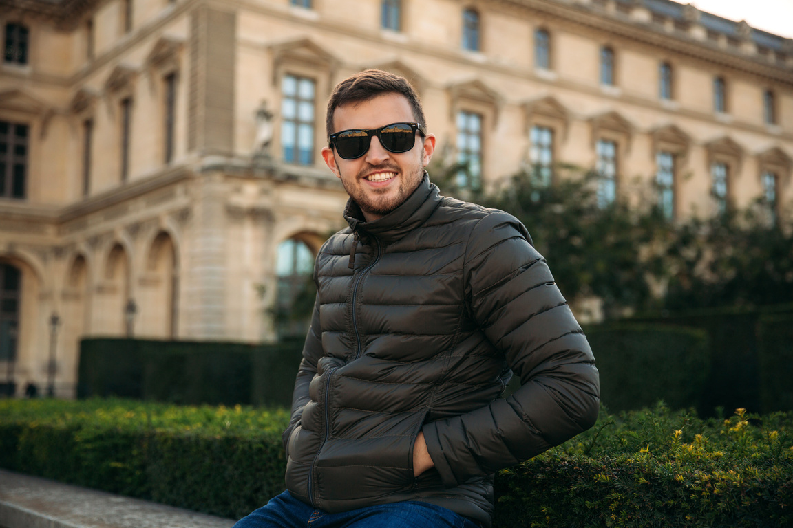 Young Man Wearing Jacket and Sunglasses Sitting Outdoors 